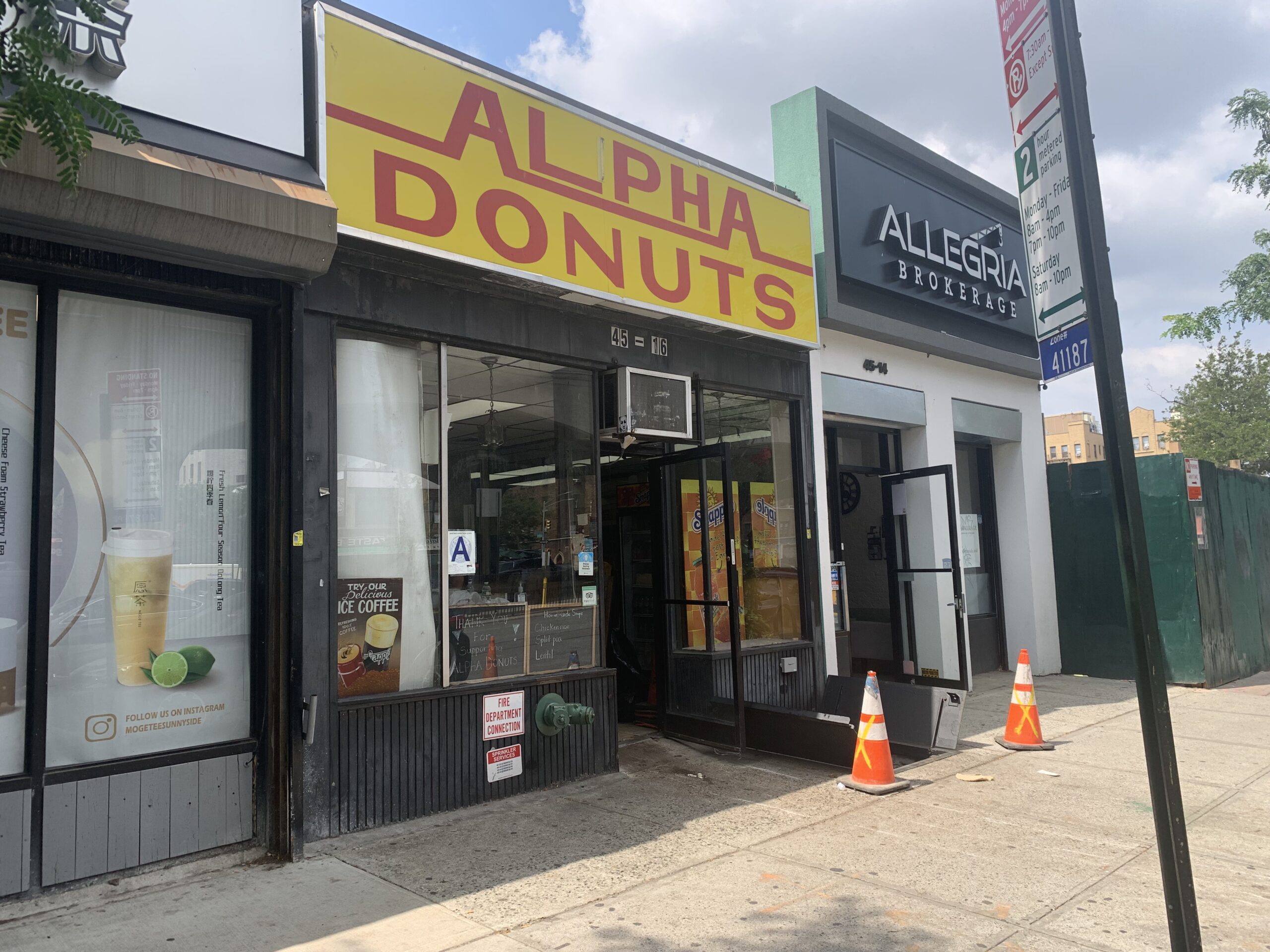 A small one-story shop sits on a street in Sunnyside, Queens, connected to two other small buildings. The shop has a bright yellow sign with the words "ALPHA DONUTS" written on it in red.