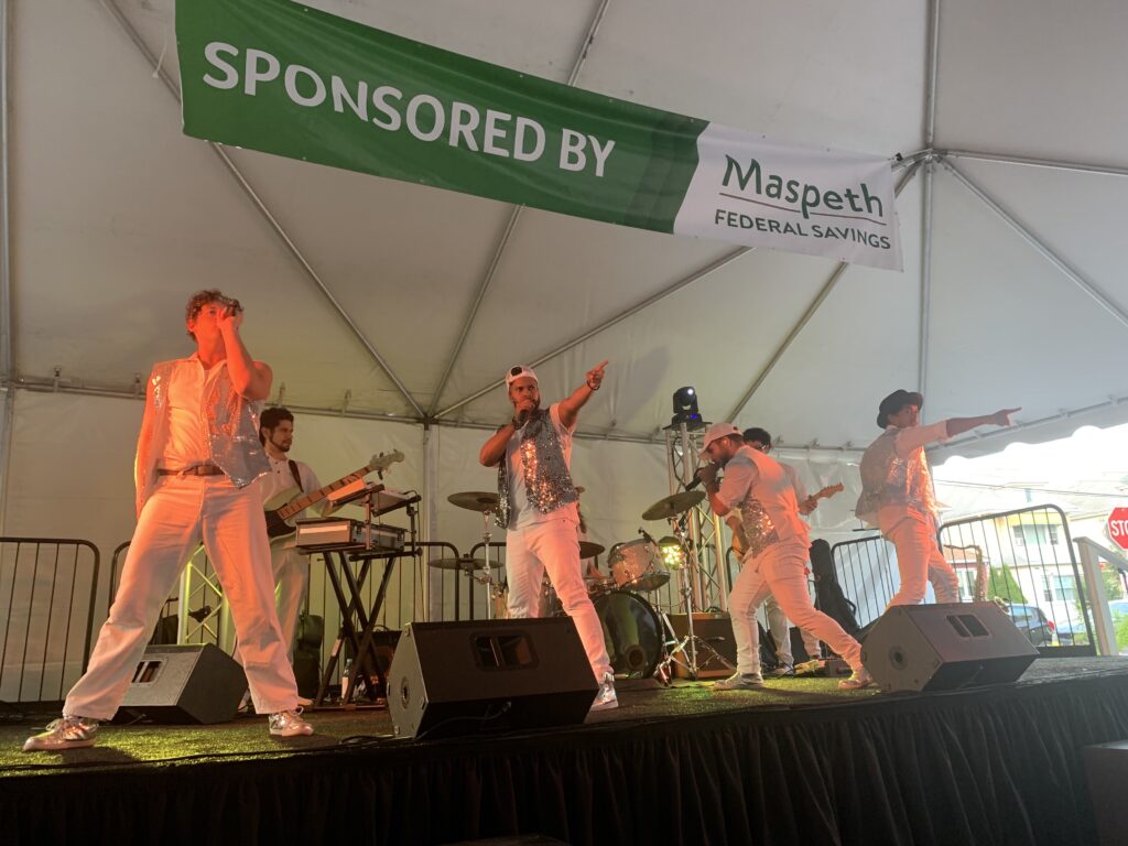 Four banmd members wearing white pants and sparkly silver vests over white t-shirts sing and dance on a black stage. They are under a white tent, and a warm orange light shines on them. A white and green sign above them reads "SPONSORED BY MASPETH FEDERAL SAVINGS."