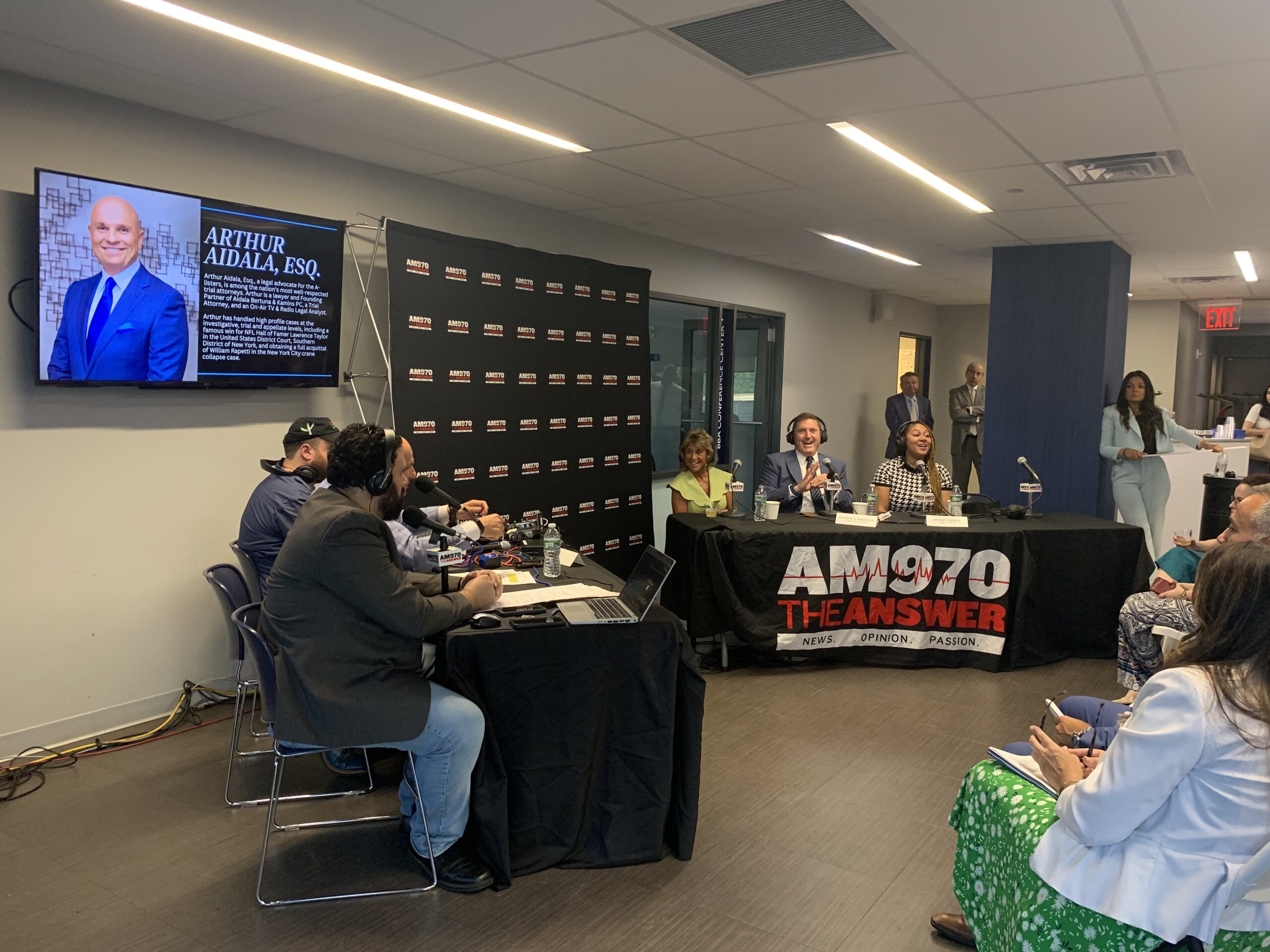 two black tables sit before a black backdrop with "AM970" written in white in a grid pattern. A tablecloth on the table to the right reads "AM970, THE ANSWER, NEWS. OPINION. PASSION." Six people sit at the tables, some wearing headphones.