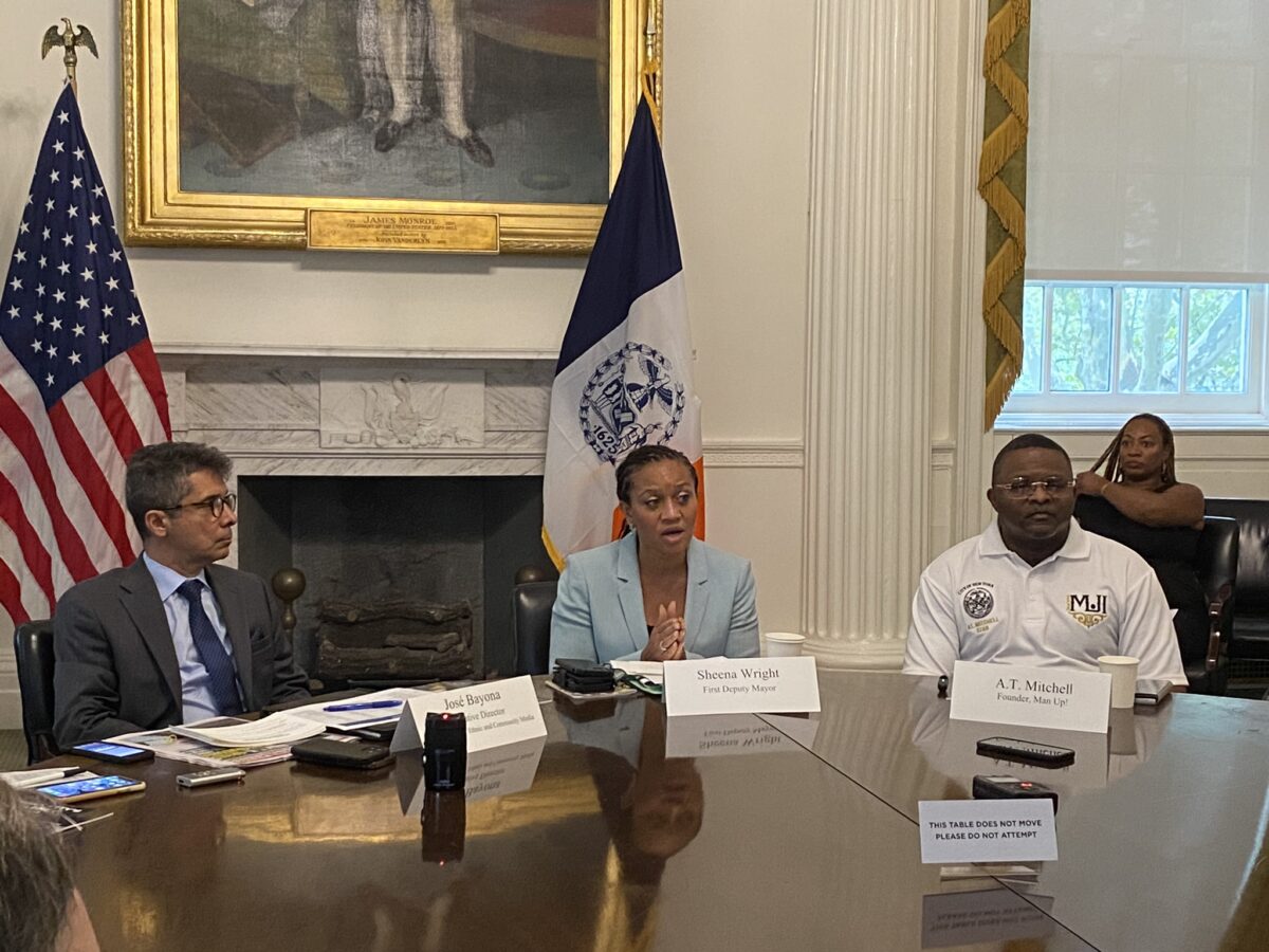 A woman in a light blue suit sits at a large wooden round table, between a man in a dark gray suit and anothe man in a whte polo shirt. Behind her, the New York State flag and the bottom of a large painting in a gold-plated frame are visible, along with a fireplace.