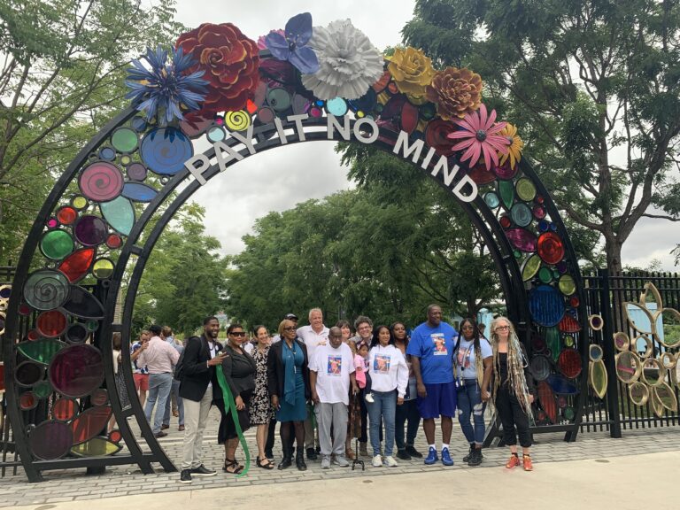 A group of around 20 people stand in front of a park gateway, posing for a photograph. The gateway is made of black metal with glass flowers and metal sculptures of flowers in different colors. Large trees with green leaves and light gray skies can be seen in the background.